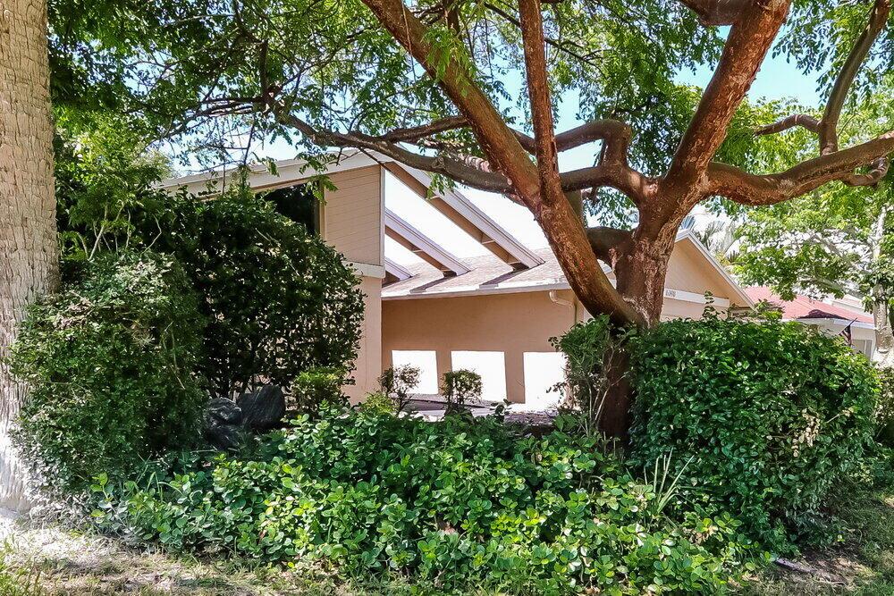 A tan house exterior is partially obscured by a large tree and dense green landscaping — BOCA RATON, FL