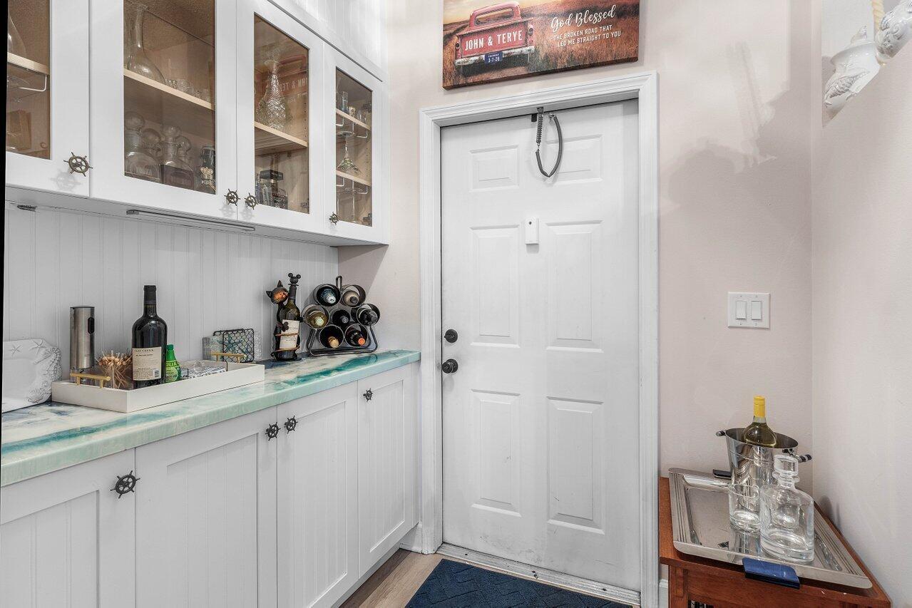 A white cabinet wet bar features a blue-toned countertop, glass-front upper cabinets, and a door — 1022 Kokomo Key Lane, Delray Beach FL 33483, DELRAY BEACH, FL