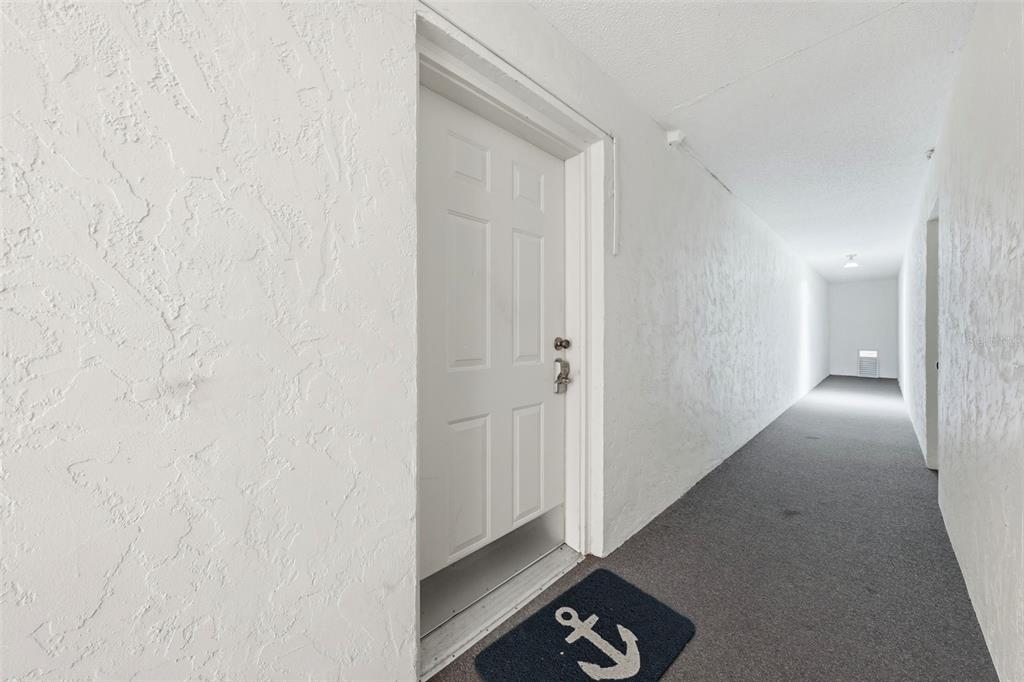 A white door with an anchor-patterned doormat sits in a carpeted, white-walled apartment hallway — 100 E OAK TERRACE DRIVE UNIT B2, LEESBURG, FL