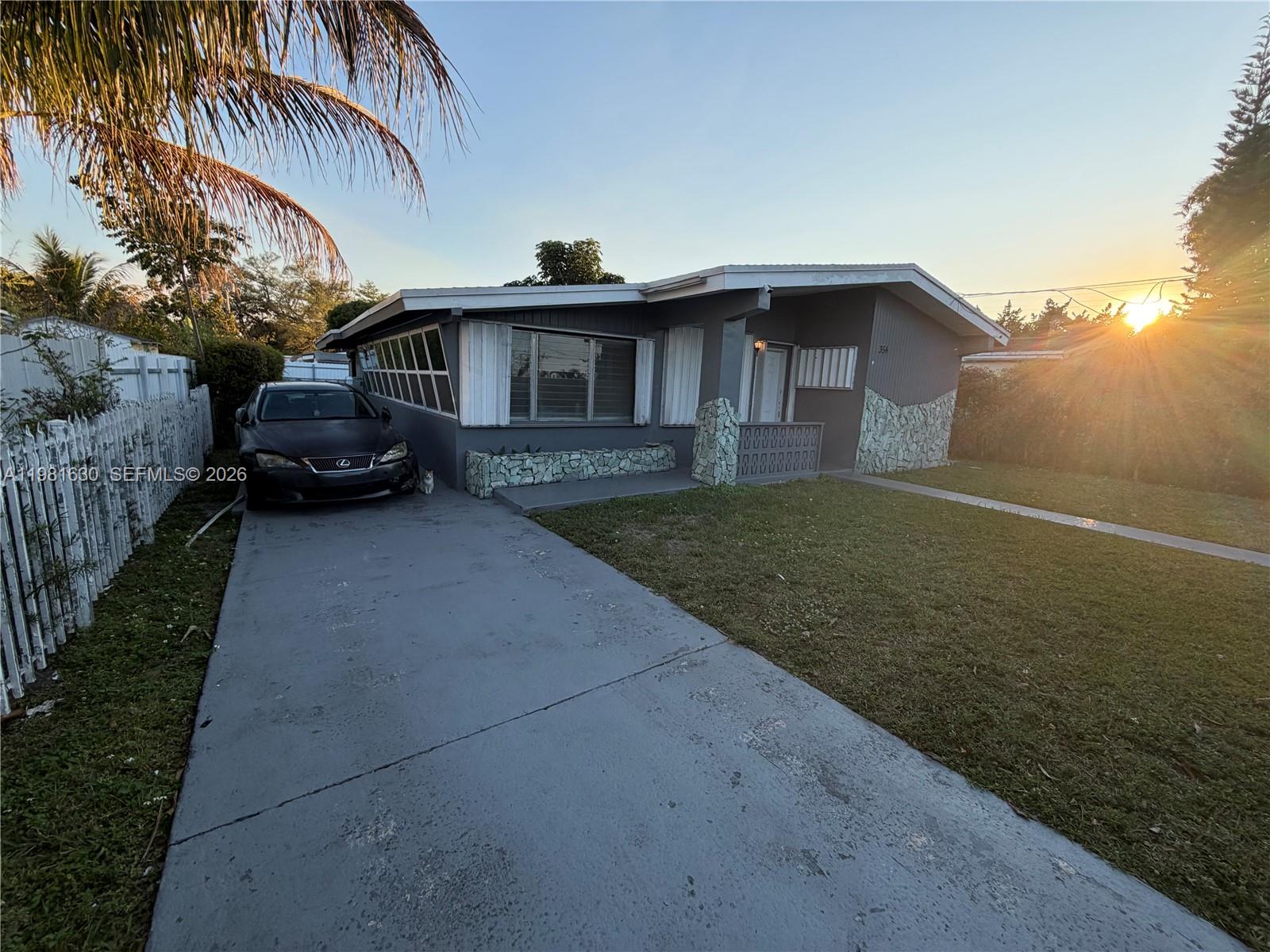 A gray single-story house with stone accents and a concrete driveway sits under a sunset — 354 NE 173rd St, North Miami Beach FL 33162, NORTH MIAMI BEACH, FL