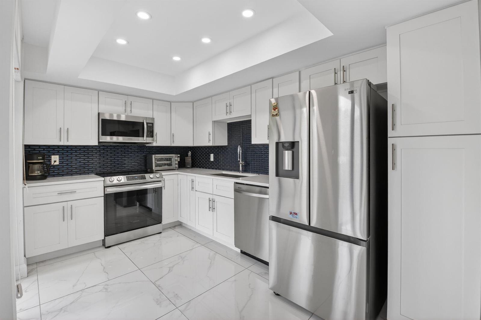 A modern kitchen features white cabinets, stainless steel appliances, a dark tile backsplash, and marble-look flooring — 6815 Willow Wood Drive # 4012, Boca Raton FL 33434, BOCA RATON, FL