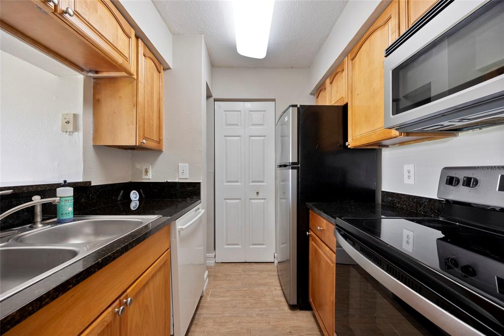 A galley kitchen features wood cabinets, black countertops, stainless steel appliances, and light wood flooring — 241 AFTON SQUARE UNIT 103, ALTAMONTE SPRINGS, FL