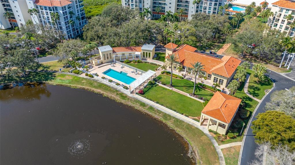 An aerial view shows a community clubhouse with a swimming pool, lawn, and adjacent lake — 4750 DOLPHIN CAY LANE S UNIT 509, ST PETERSBURG, FL