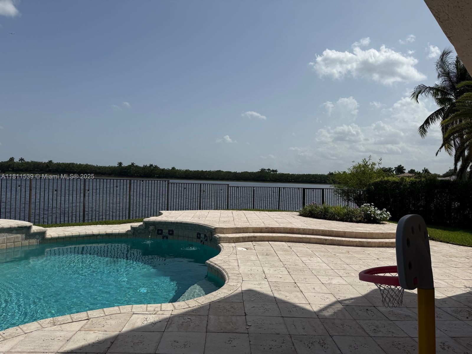 A swimming pool with a stone deck overlooks a lake behind a metal fence — 1418 Lantana Dr, Weston FL 33326, WESTON, FL