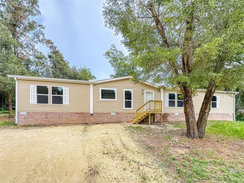 A tan manufactured home with a stone-patterned base sits on a dirt lot with trees — 12841 SE 103RD TERRACE, BELLEVIEW, FL