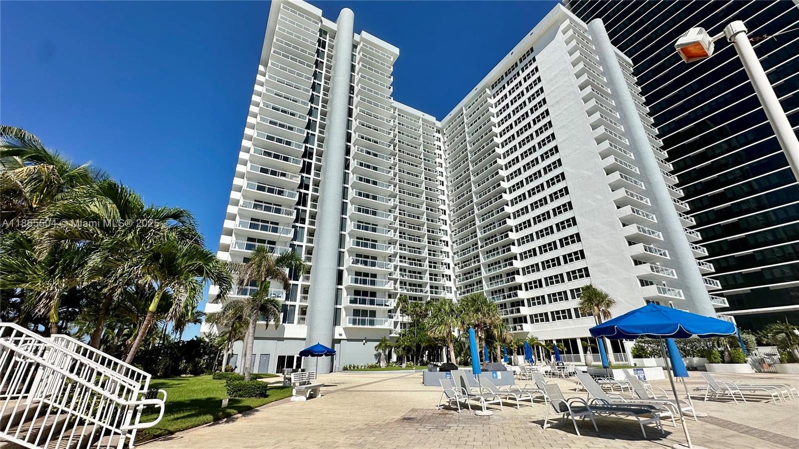 A tall white residential building features numerous balconies overlooking a paved pool deck with umbrellas — 2030 S Ocean Dr # 1022, Hallandale Beach FL 33009, HALLANDALE BEACH, FL