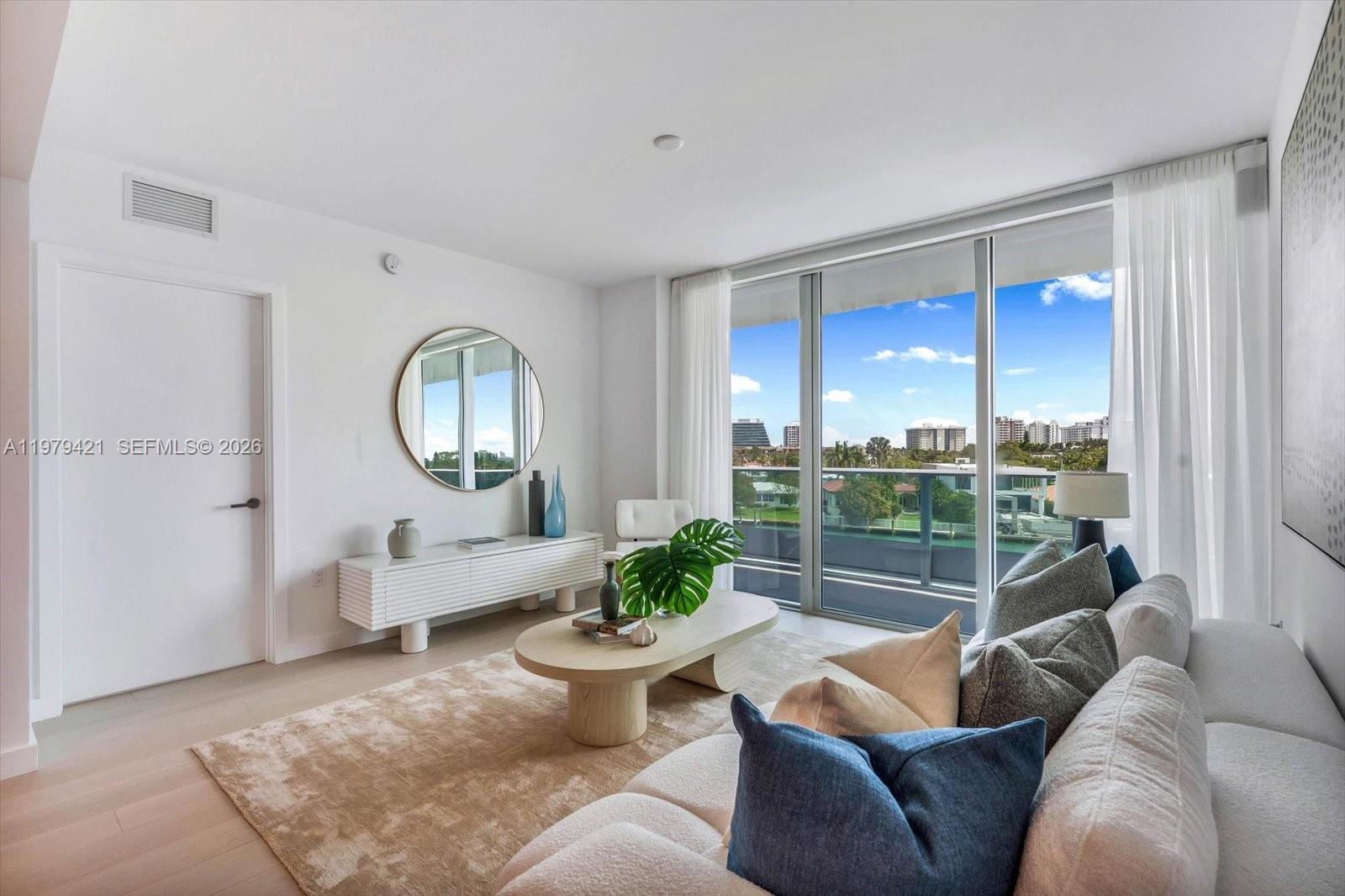A modern living room features a white console, round mirror, and balcony with water views — 9201 E Bay Harbor Dr # 503, Bal Harbour FL 33154, BAL HARBOUR, FL