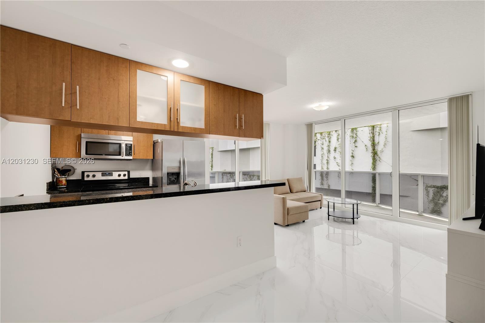 A kitchen with wood cabinets and black countertops opens into a living area with balcony — 601 NE 23rd St # 706, Miami FL 33137, MIAMI, FL