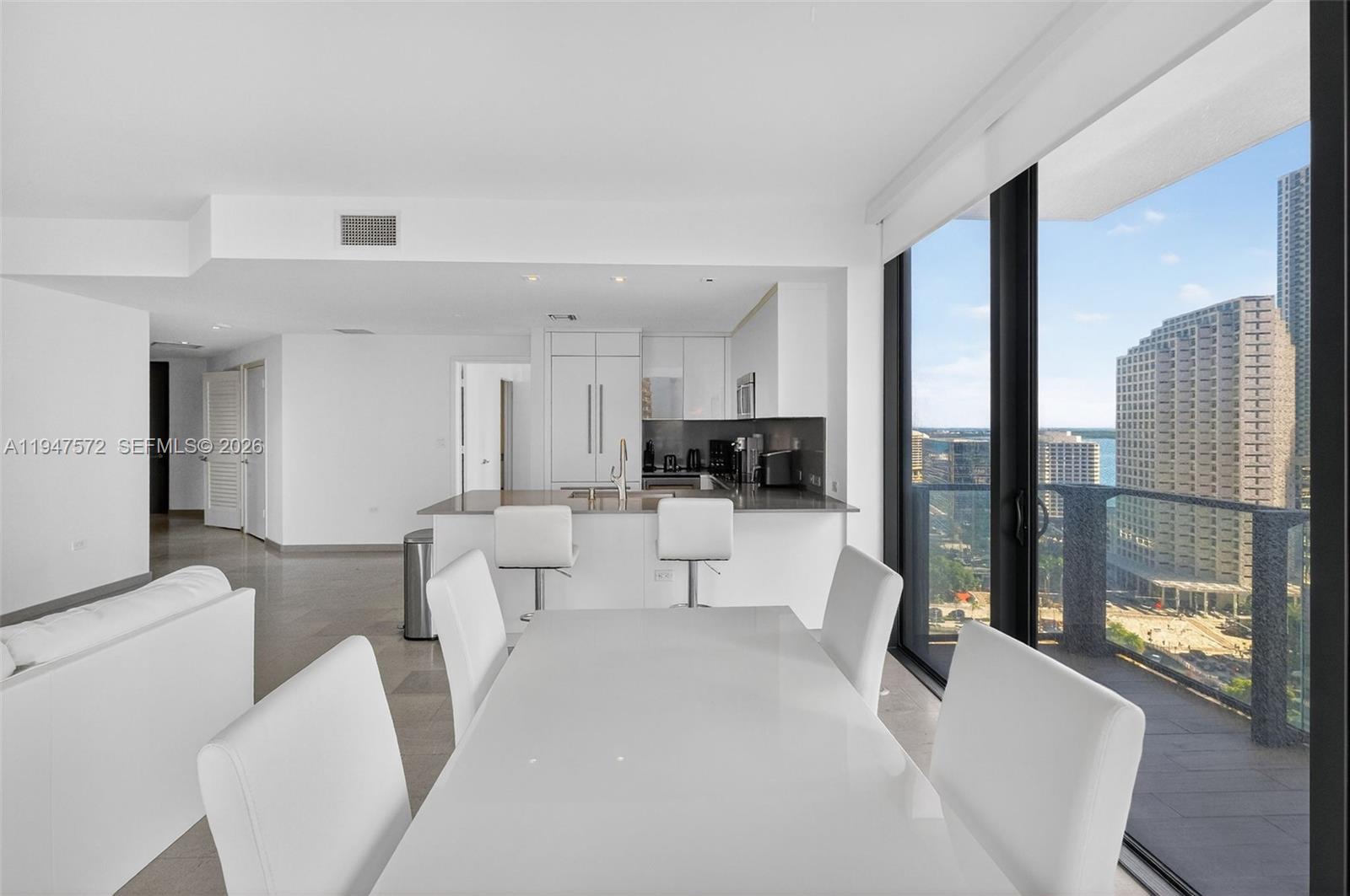 A modern white dining area and kitchen overlook a city skyline through floor-to-ceiling windows — 68 SE 6th St # 1702, Miami FL 33131, MIAMI, FL
