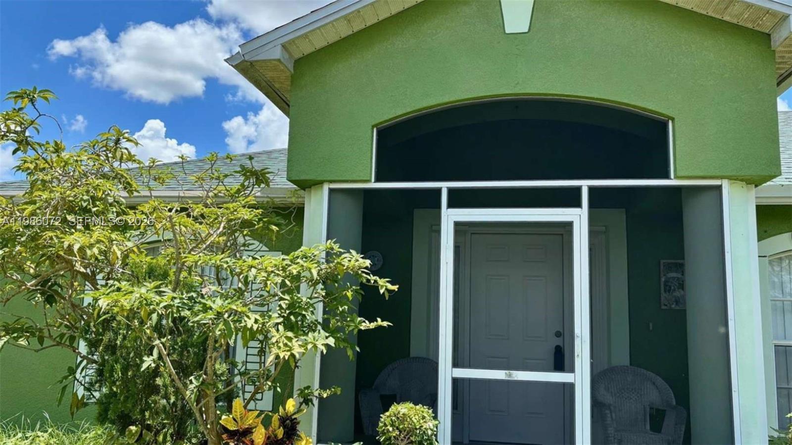 A green house exterior features a screened-in front porch with two wicker chairs — 1122 MCKINLEY AVE, Lehigh Acres FL 33972, LEHIGH ACRES, FL