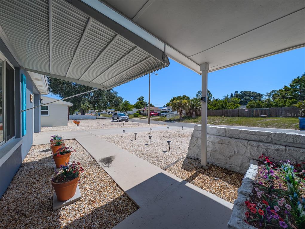 A covered concrete patio features potted flowers, a stone retaining wall, and a gravel yard — 3702 KINGSBURY, HOLIDAY, FL