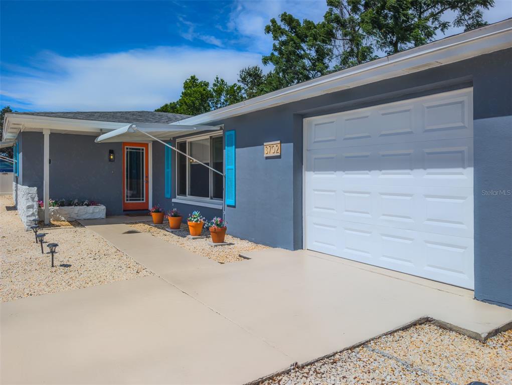 A gray house exterior features a white garage door, an orange front door, and landscaping — 3702 KINGSBURY, HOLIDAY, FL