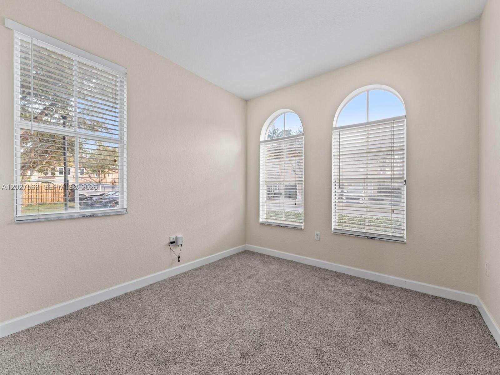 An empty room features beige walls, gray carpeting, and three windows with white blinds — 1498 SE 25th Ave, Homestead FL 33035, HOMESTEAD, FL