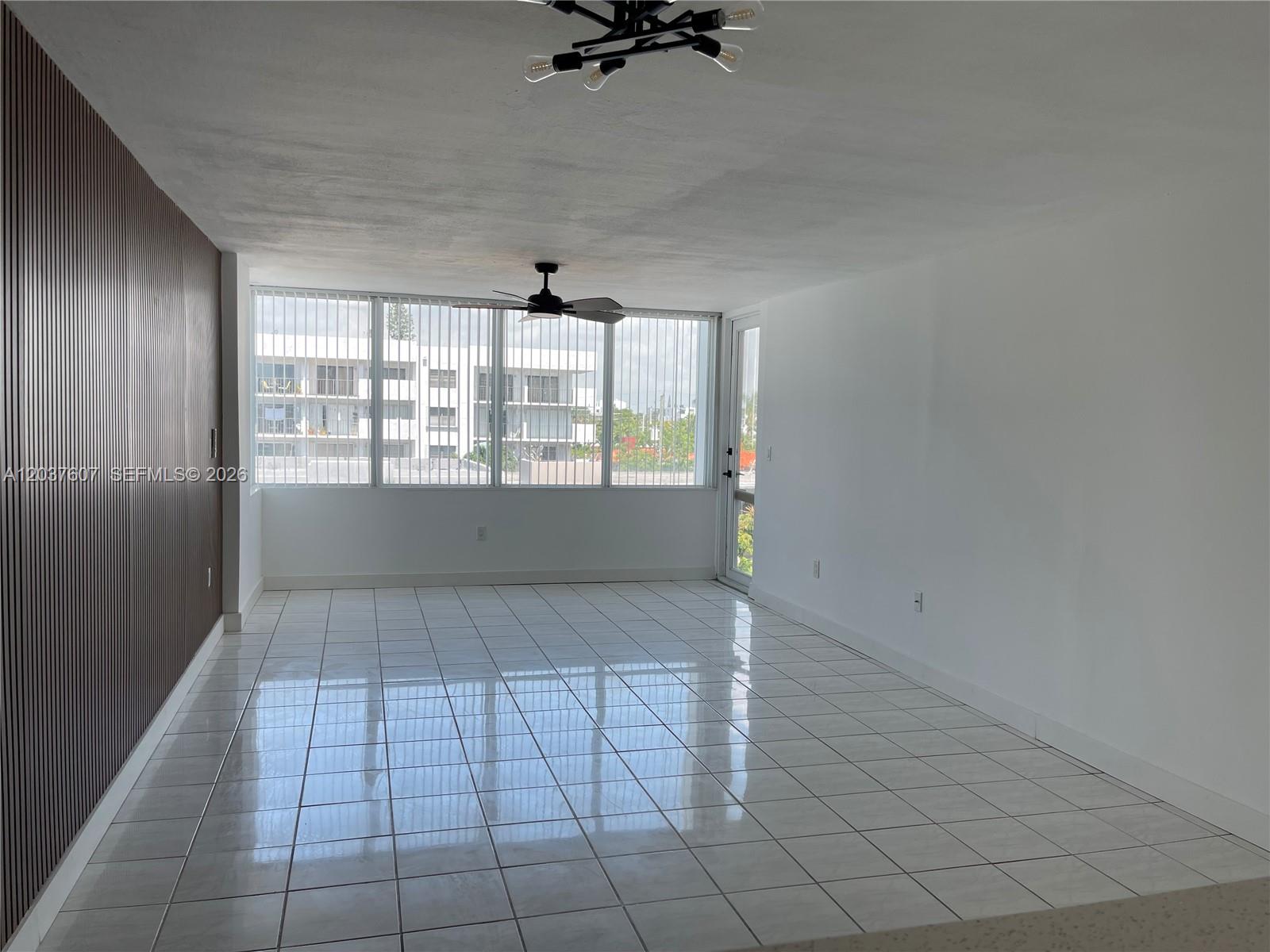 An empty room features white tile flooring, a slatted wood accent wall, and large windows — 8233 Harding Ave # 408, Miami Beach FL 33141, MIAMI BEACH, FL