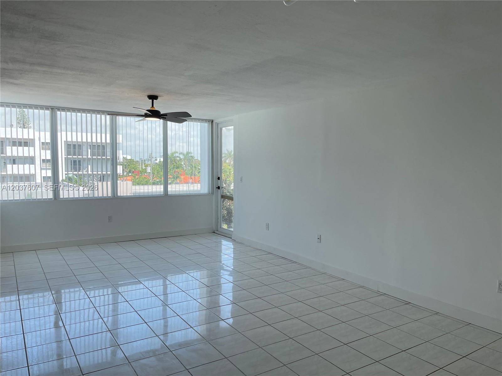 An empty room features white tiled floors, a ceiling fan, and large windows with blinds — 8233 Harding Ave # 408, Miami Beach FL 33141, MIAMI BEACH, FL