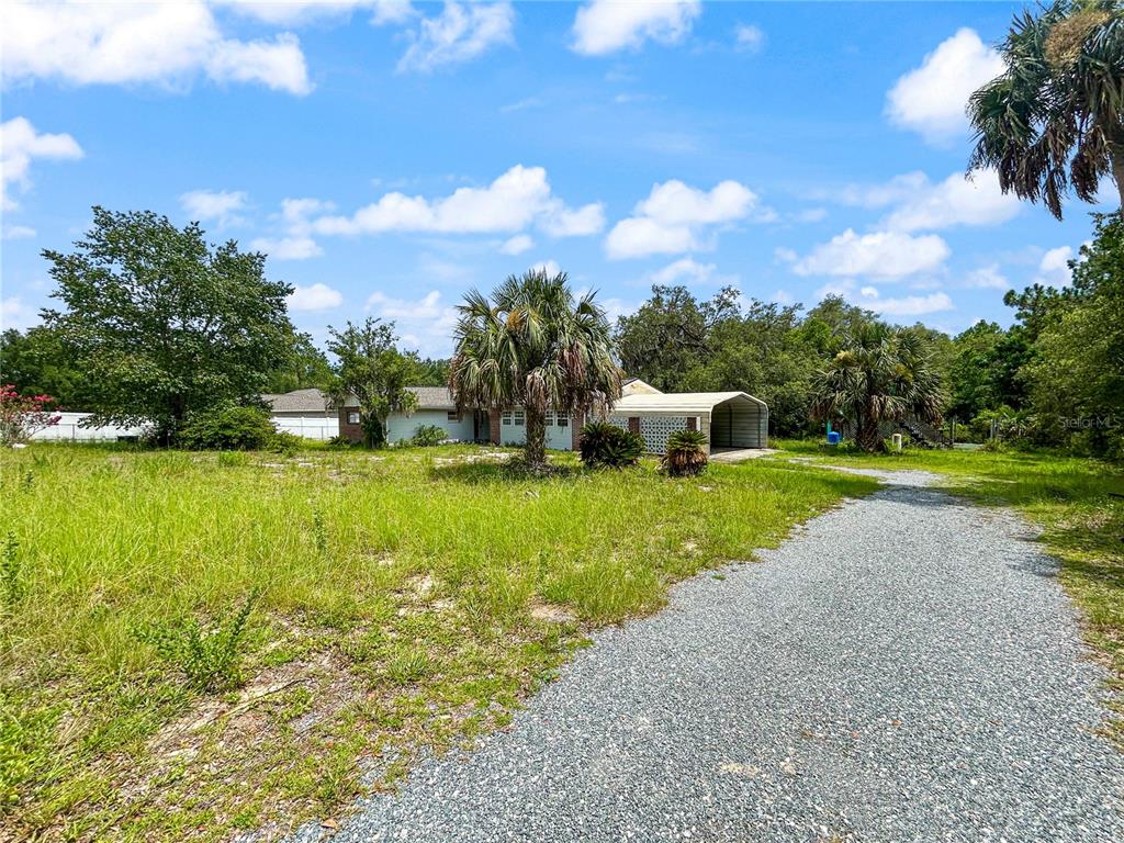 A gravel driveway leads to a single-story house with a metal carport and palm trees — 23162 SW NEPTUNE BOULEVARD, DUNNELLON, FL
