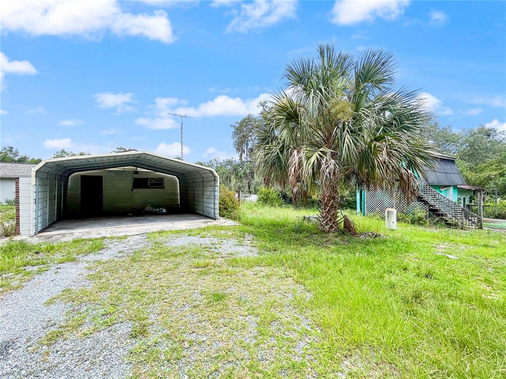 A metal carport sits on a gravel driveway next to a palm tree and house — 23162 SW NEPTUNE BOULEVARD, DUNNELLON, FL