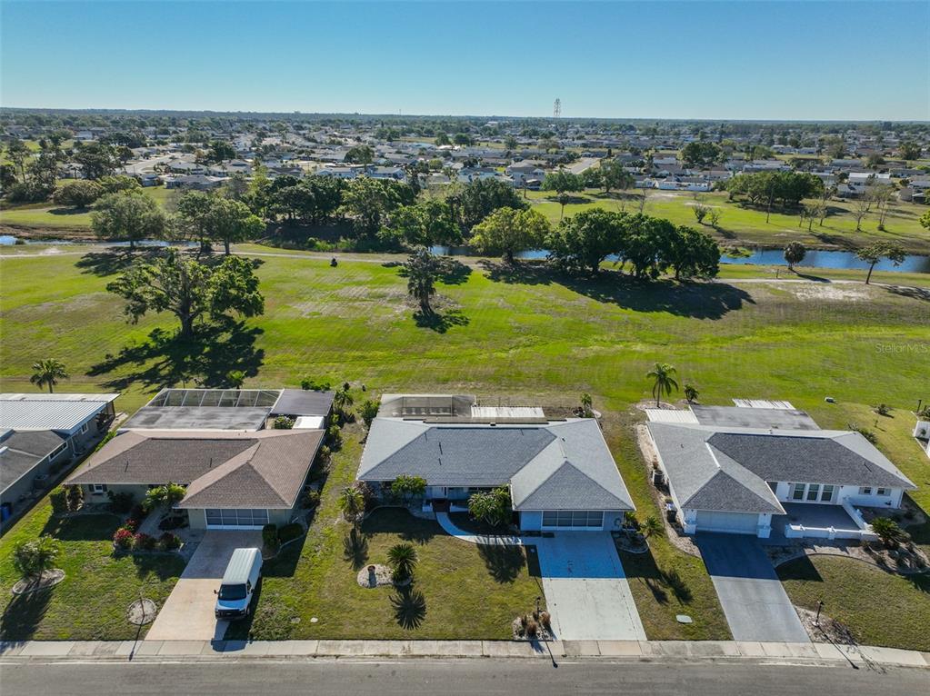 An aerial view shows three suburban houses with backyards facing a large grassy field and pond — 672 ALLEGHENY DRIVE, SUN CITY CENTER, FL
