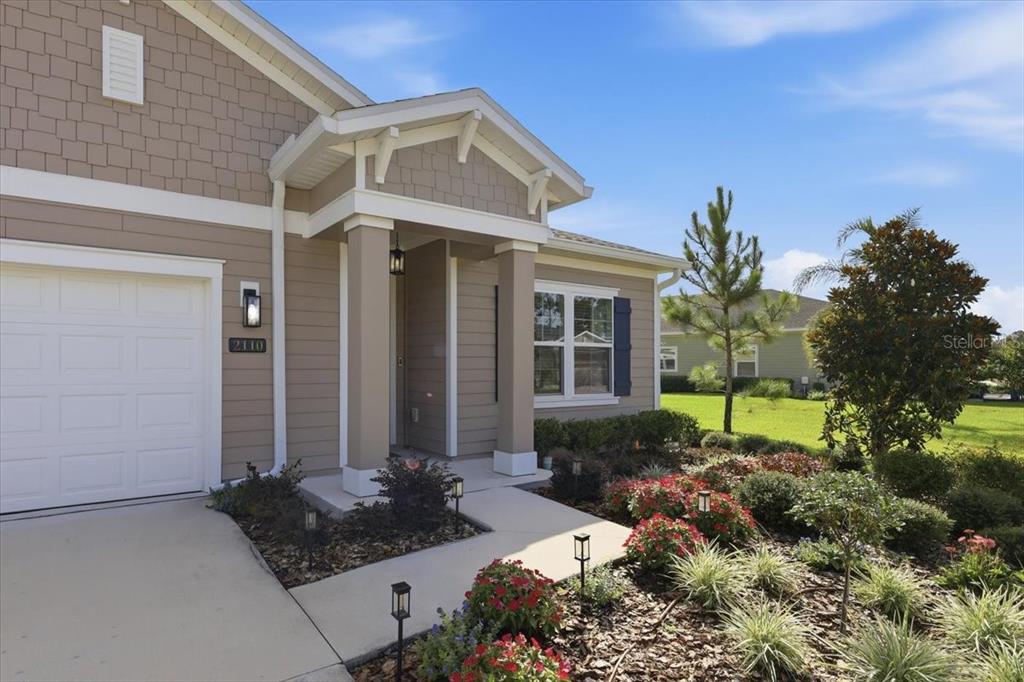 A tan house exterior features a white garage door, a covered entryway, and landscaped gardens — 2110 RAGLAN CIRCLE, ORMOND BEACH, FL