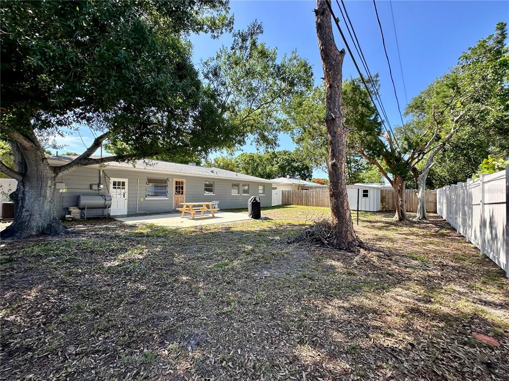 A backyard with a gray house, a concrete patio, a picnic table, and mature trees — 4917 99TH STREET N, ST PETERSBURG, FL