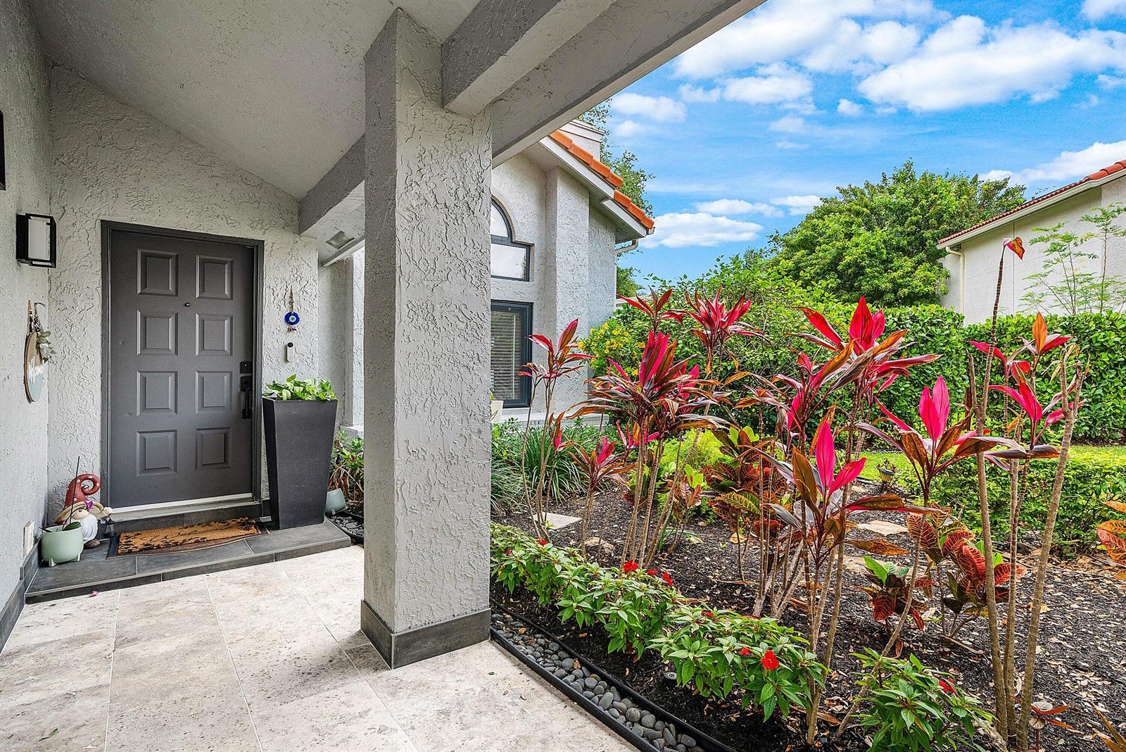 A covered entryway features a gray front door, tiled flooring, and a landscaped garden bed — 20797 Cabrillo Way, Boca Raton FL 33428, BOCA RATON, FL