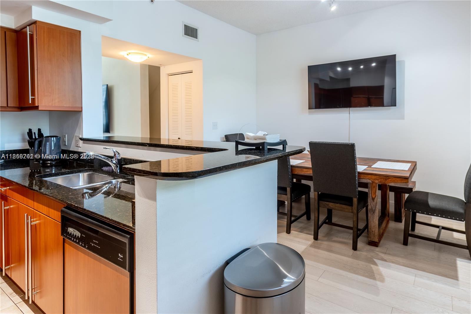 A kitchen with wooden cabinets and granite countertops overlooks a dining area with a television — 2069 S Ocean Dr, Hallandale Beach FL 33009, HALLANDALE BEACH, FL