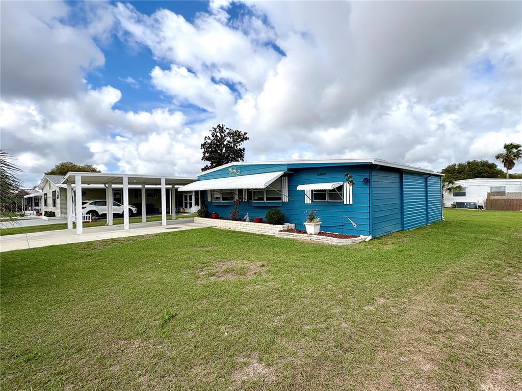 A blue mobile home with a white carport sits on a grassy lot under clouds — 7481 FIRST CIRCLE DRIVE, BROOKSVILLE, FL