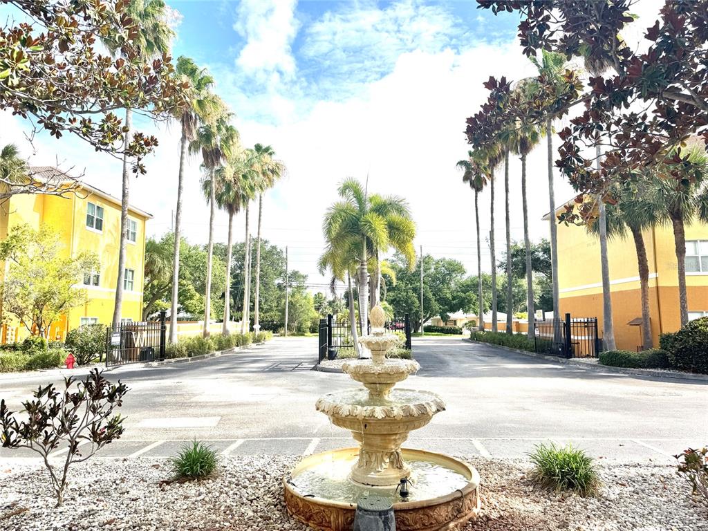 A stone fountain sits in the foreground of a paved entrance between two yellow buildings — 10764 70TH AVENUE UNIT #1201, SEMINOLE, FL