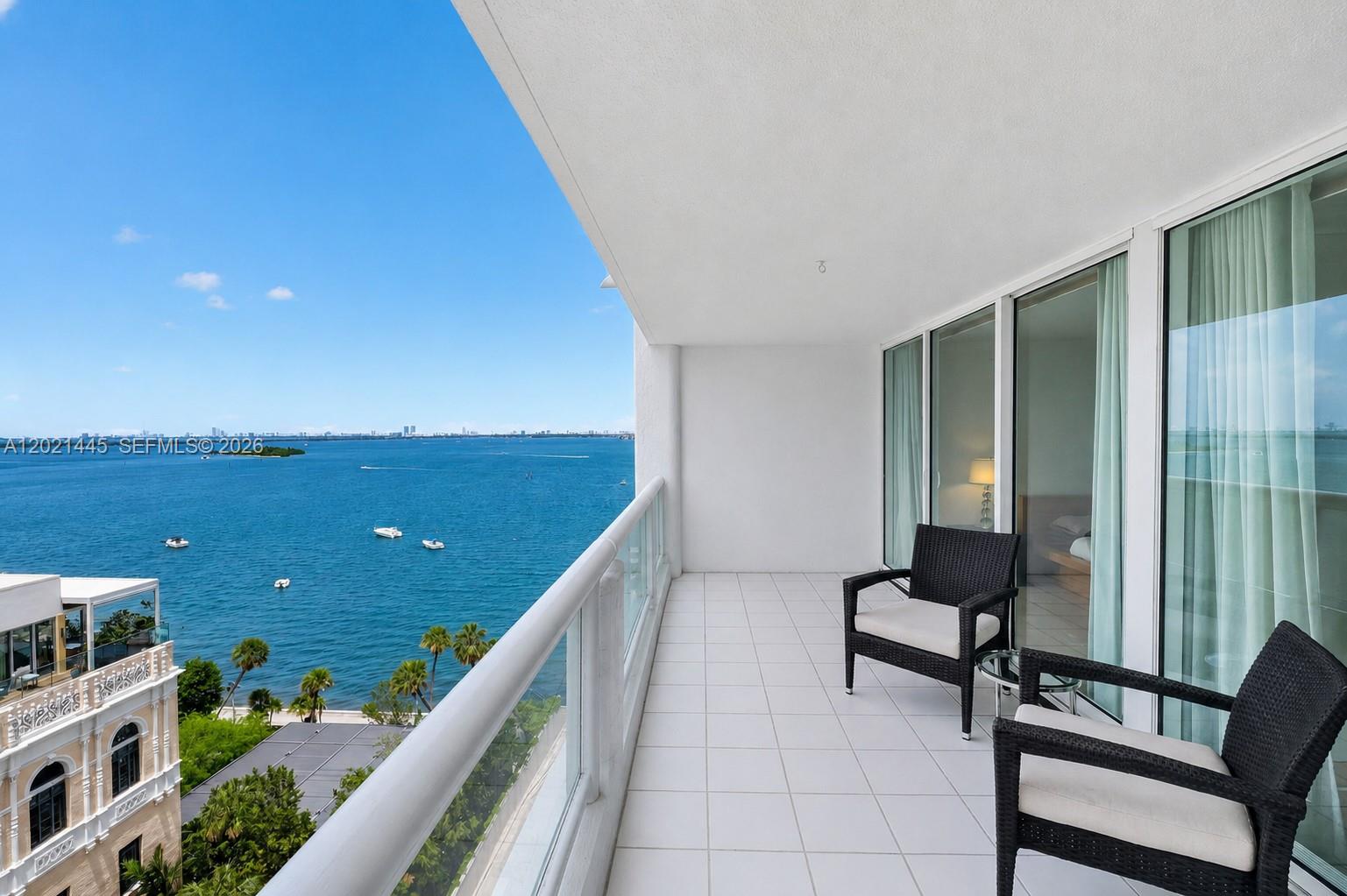 A balcony with white tile flooring and two wicker chairs overlooks a large body of water — 1717 N Bayshore Dr # 1437, Miami FL 33132, MIAMI, FL