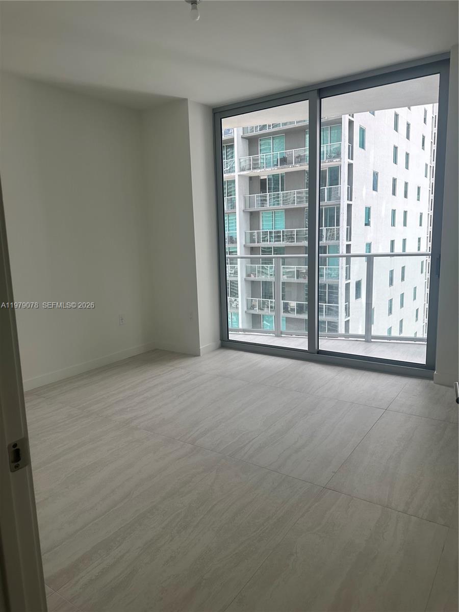 A room with light-colored tile flooring and a large sliding glass door overlooking buildings — 77 SE 5th # 2514, Miami FL 33131, MIAMI, FL