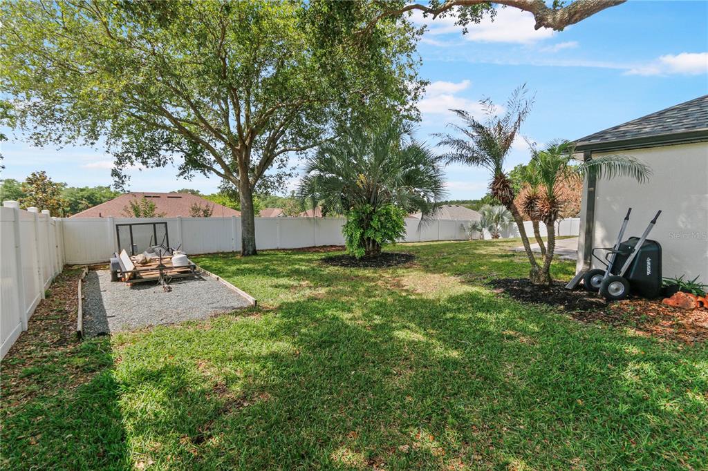 A grassy backyard features a white vinyl fence, a gravel pad with a trailer, and trees — 10814 VINEYARD COURT, CLERMONT, FL