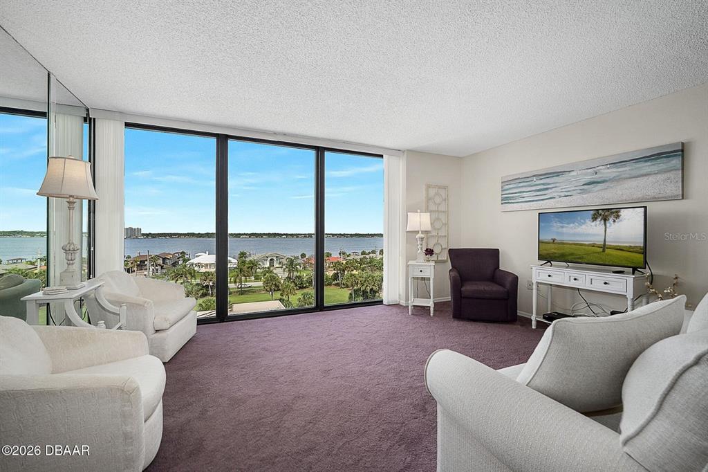 A living room features purple carpeting, white furniture, and floor-to-ceiling windows overlooking a waterfront view — 1 OCEANS WEST BOULEVARD UNIT 6A6, DAYTONA BEACH, FL