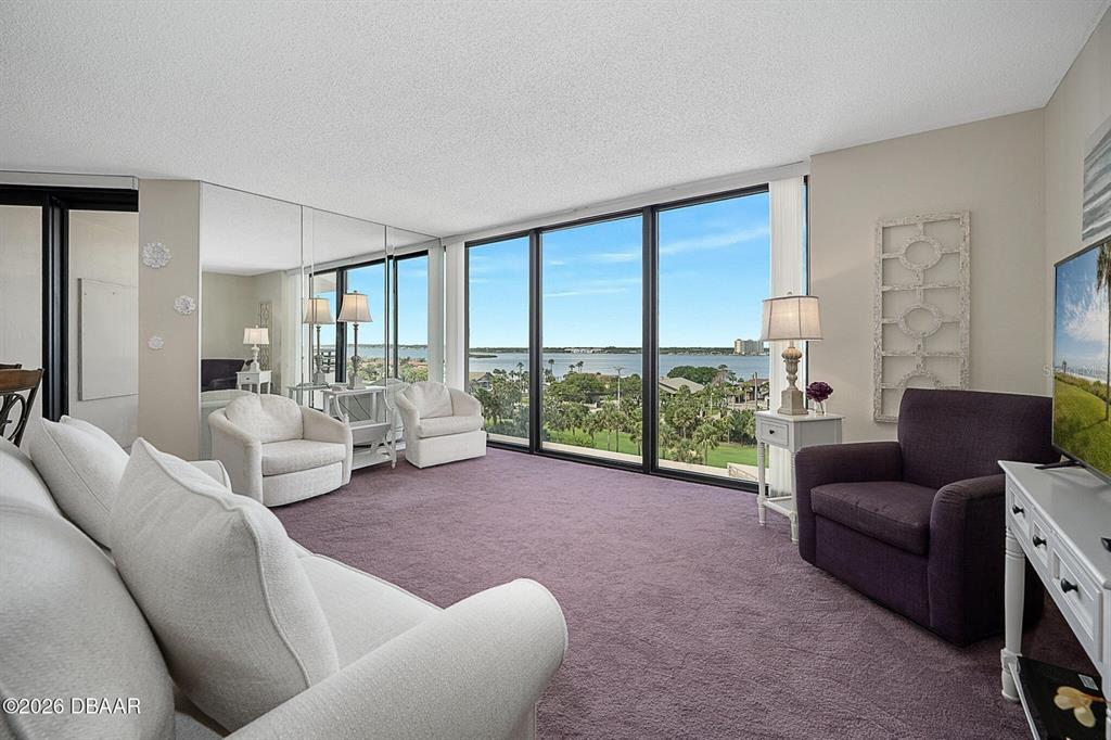 A living room features purple carpet, white furniture, mirrored walls, and large windows overlooking water — 1 OCEANS WEST BOULEVARD UNIT 6A6, DAYTONA BEACH, FL