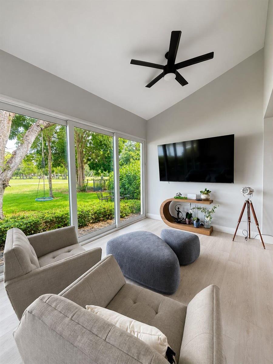 A modern living room features two armchairs, grey ottomans, a wall-mounted television, and large windows — 4103 Carriage Drive # H4, Pompano Beach FL 33069, POMPANO BEACH, FL