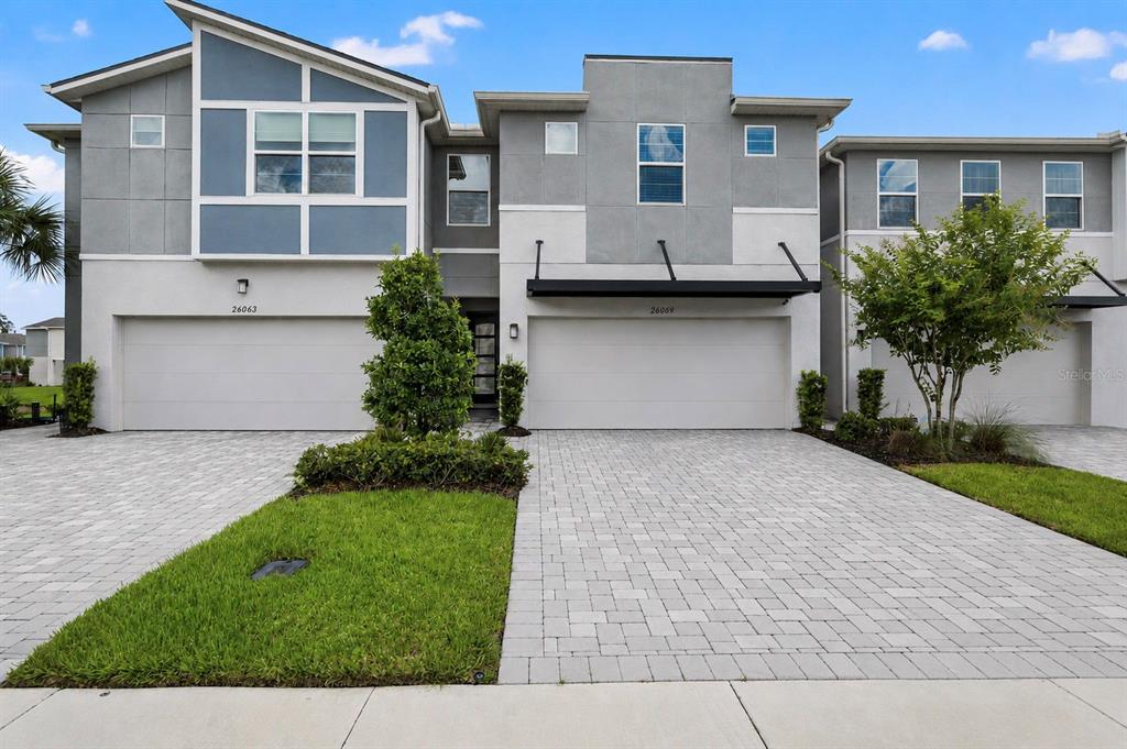 A two-story duplex with gray stucco siding, two garage doors, and a paved driveway — 26069 WOVEN WICKER BEND, LUTZ, FL