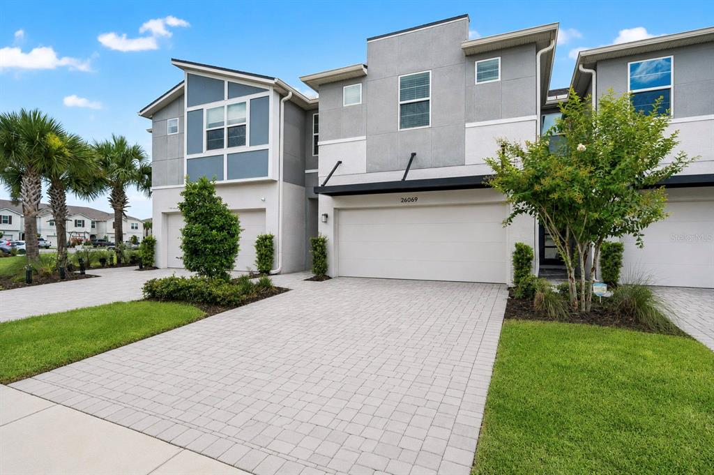 A two-story townhouse exterior features a paved driveway, a two-car garage, and palm trees — 26069 WOVEN WICKER BEND, LUTZ, FL