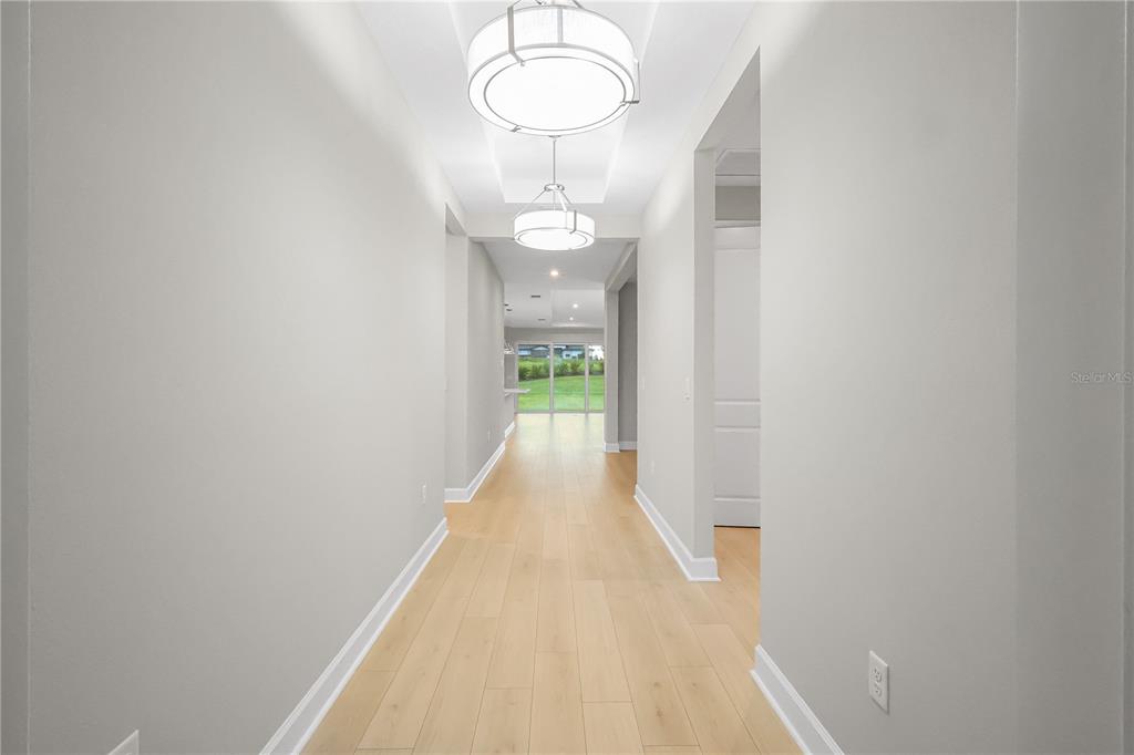 A long, light-filled hallway features light wood flooring, white walls, and two modern drum chandeliers — 1620 BELLAGIO LOOP, TAVARES, FL