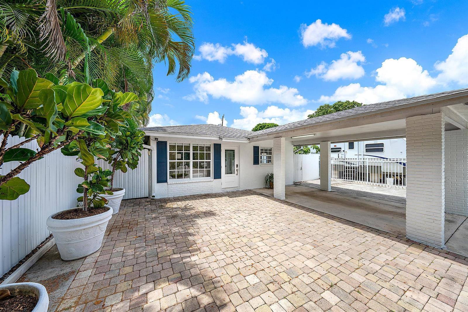 A white house exterior features a brick-paved driveway, a covered carport, and large potted plants — 904 Ortega Road, West Palm Beach FL 33405, WEST PALM BEACH, FL