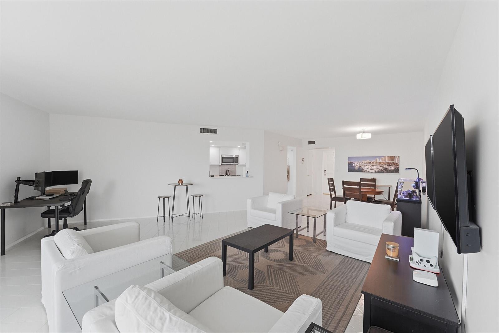 A bright, open-concept living room features white furniture, a dark coffee table, and a desk area — 2681 S Course Drive # 808, Pompano Beach FL 33069, POMPANO BEACH, FL