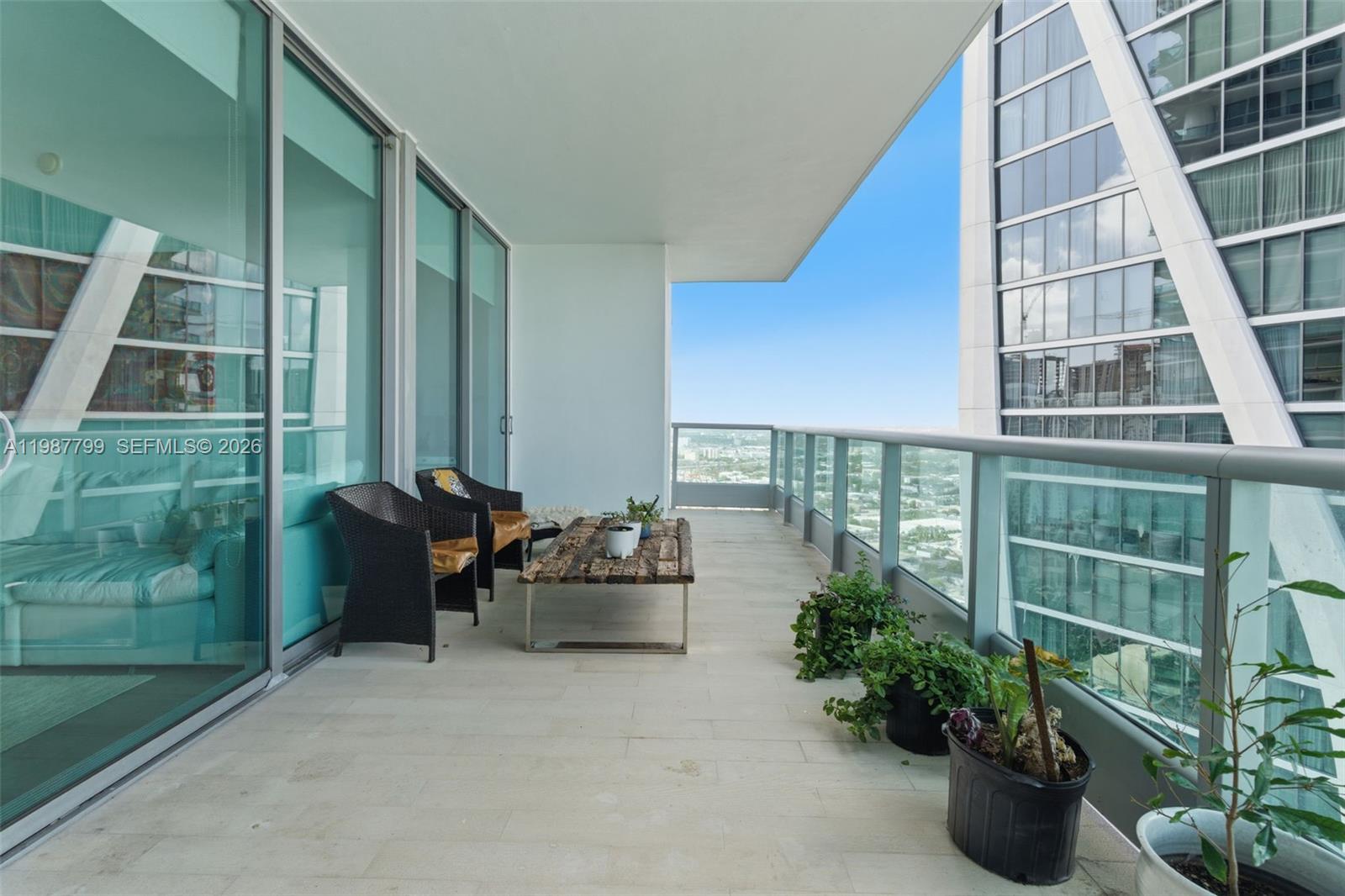 A high-rise balcony features a wooden coffee table, two wicker chairs, and several potted plants — 900 Biscayne Blvd # 4308, Miami FL 33132, MIAMI, FL