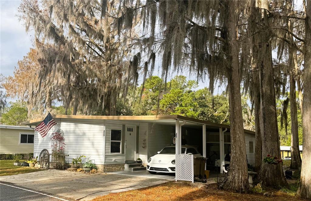 A single-story mobile home with a carport and an American flag under large mossy trees — 107 ROYAL PALM DRIVE, LEESBURG, FL