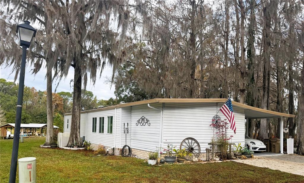 A white mobile home with a carport and an American flag stands among mossy trees — 107 ROYAL PALM DRIVE, LEESBURG, FL