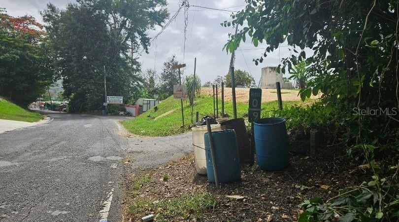 A paved road leads past a grassy embankment with three large plastic barrels — PR-633 KM 0.9 BO. BARAHONA WD,, MOROVIS, PR