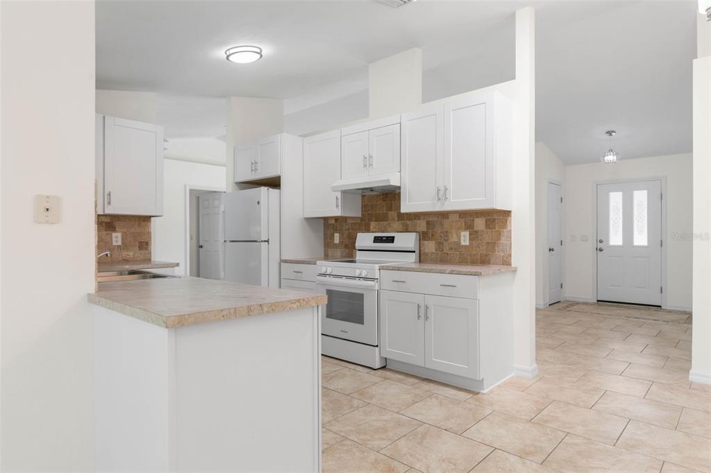 A kitchen with white cabinets, a tiled backsplash, and light-colored countertops opens to an entryway — 23 PENNFIELD LANE, PALM COAST, FL