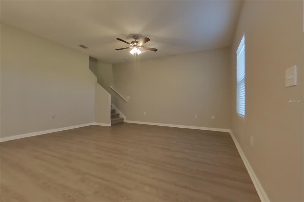 An empty living room features light-colored walls, wood-look flooring, a ceiling fan, and stairs — 15619 MERLIN AVENUE, MASCOTTE, FL