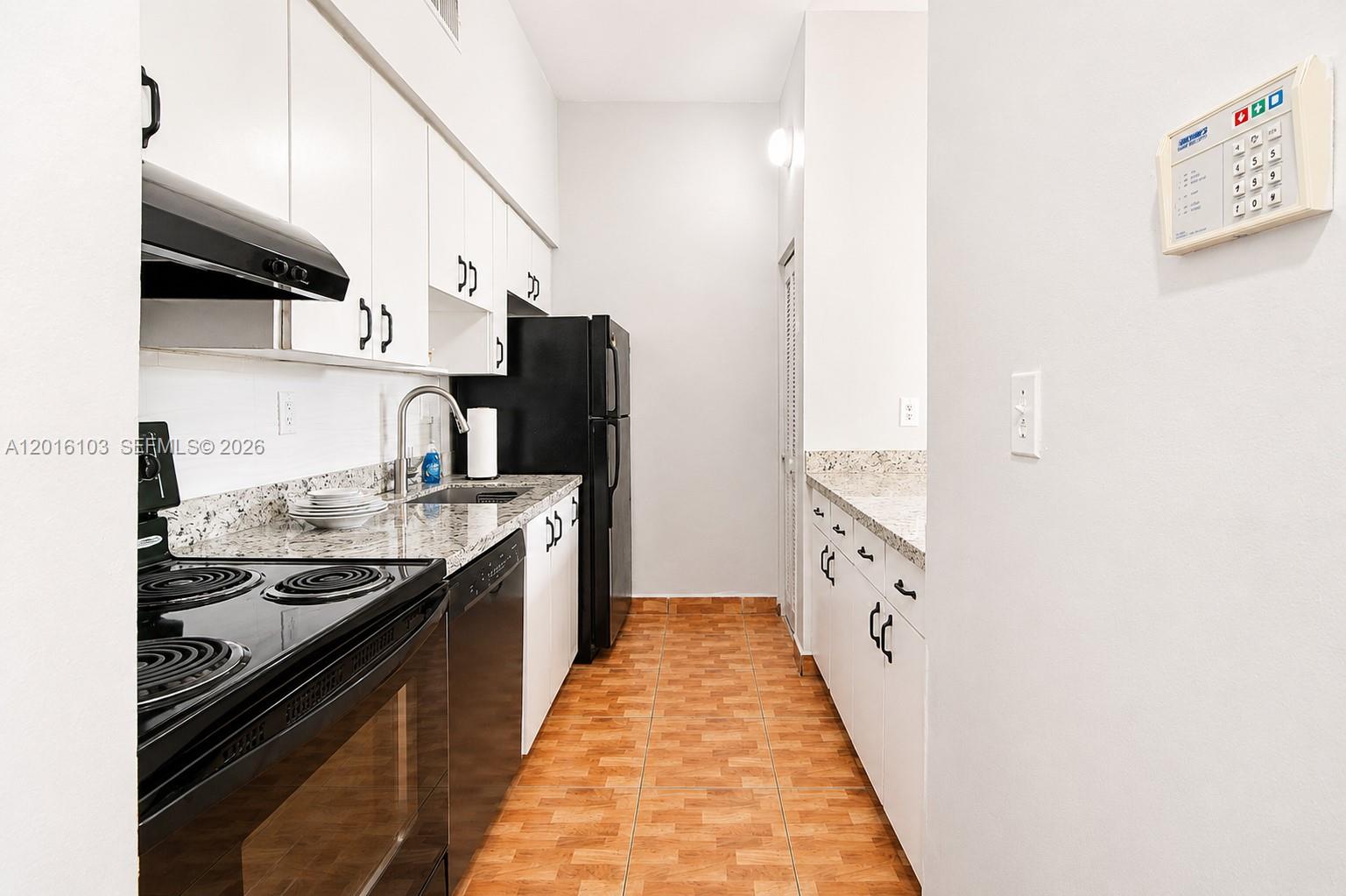 A narrow galley kitchen features white cabinets, black appliances, granite countertops, and wood-look tile flooring — 7400 W 20th Ave # 312, Hialeah FL 33016, HIALEAH, FL