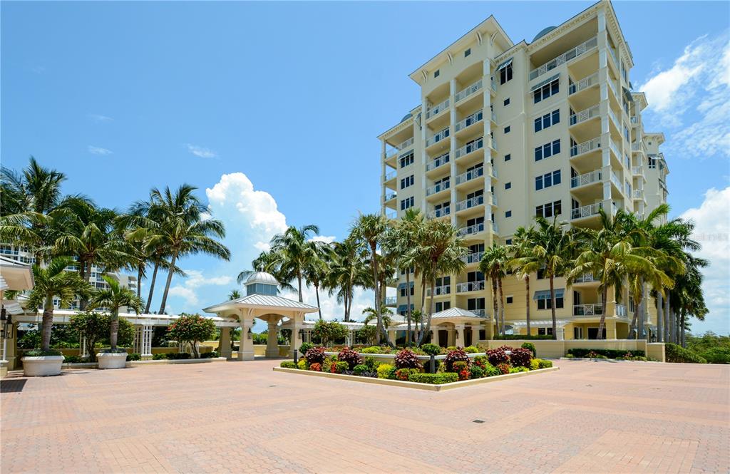 A multi-story beige residential building stands behind a paved courtyard with palm trees — 2050 BENJAMIN FRANKLIN DRIVE UNIT B704, SARASOTA, FL