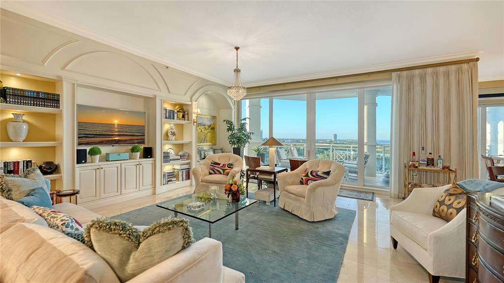 A spacious living room features built-in shelving, a large television, and floor-to-ceiling windows with views — 2050 BENJAMIN FRANKLIN DRIVE UNIT B704, SARASOTA, FL