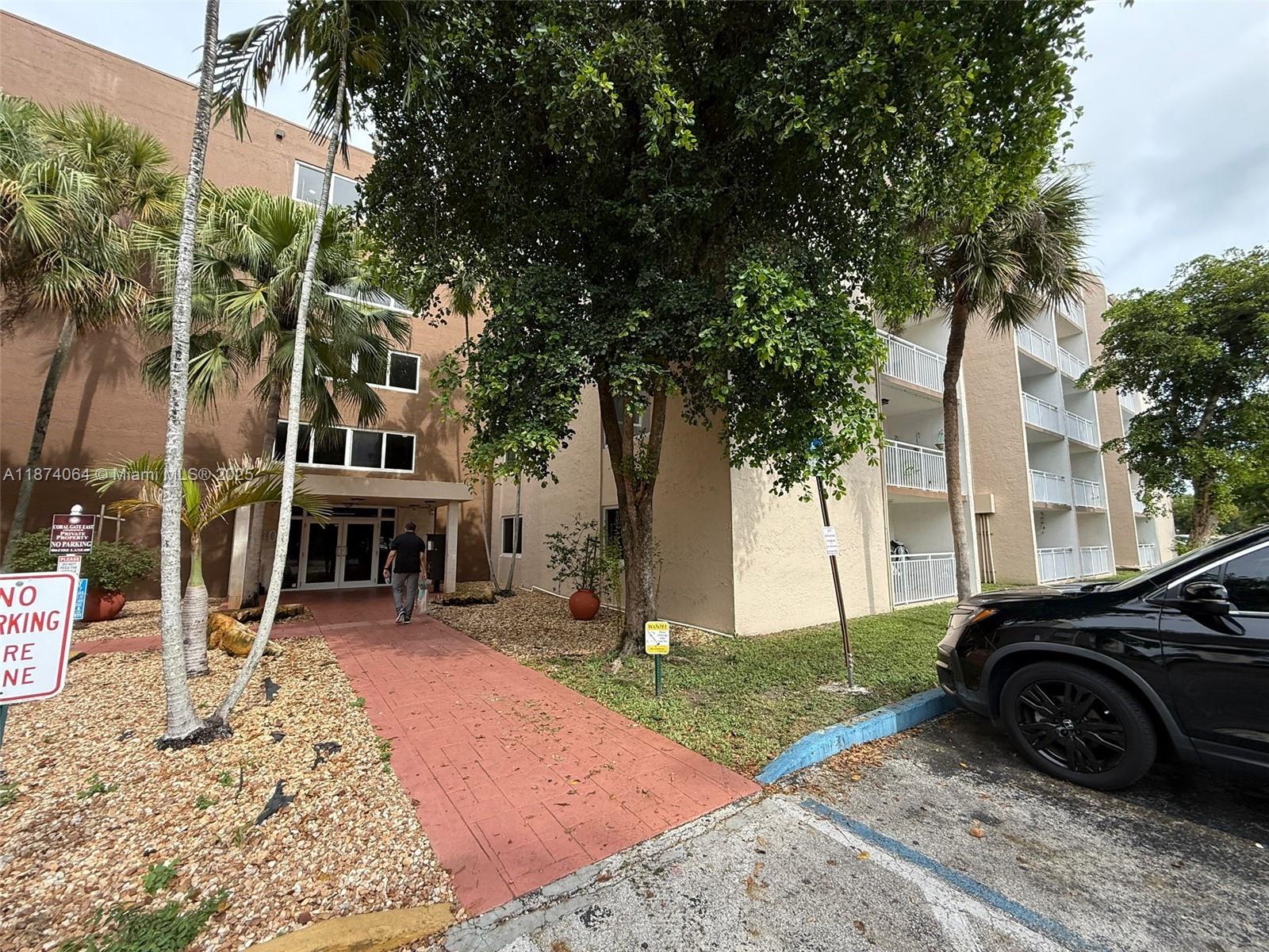 A paved walkway leads to the entrance of a multi-story apartment building with palm trees — 6940 Miami Gardens Dr # 122, Hialeah FL 33015, HIALEAH, FL
