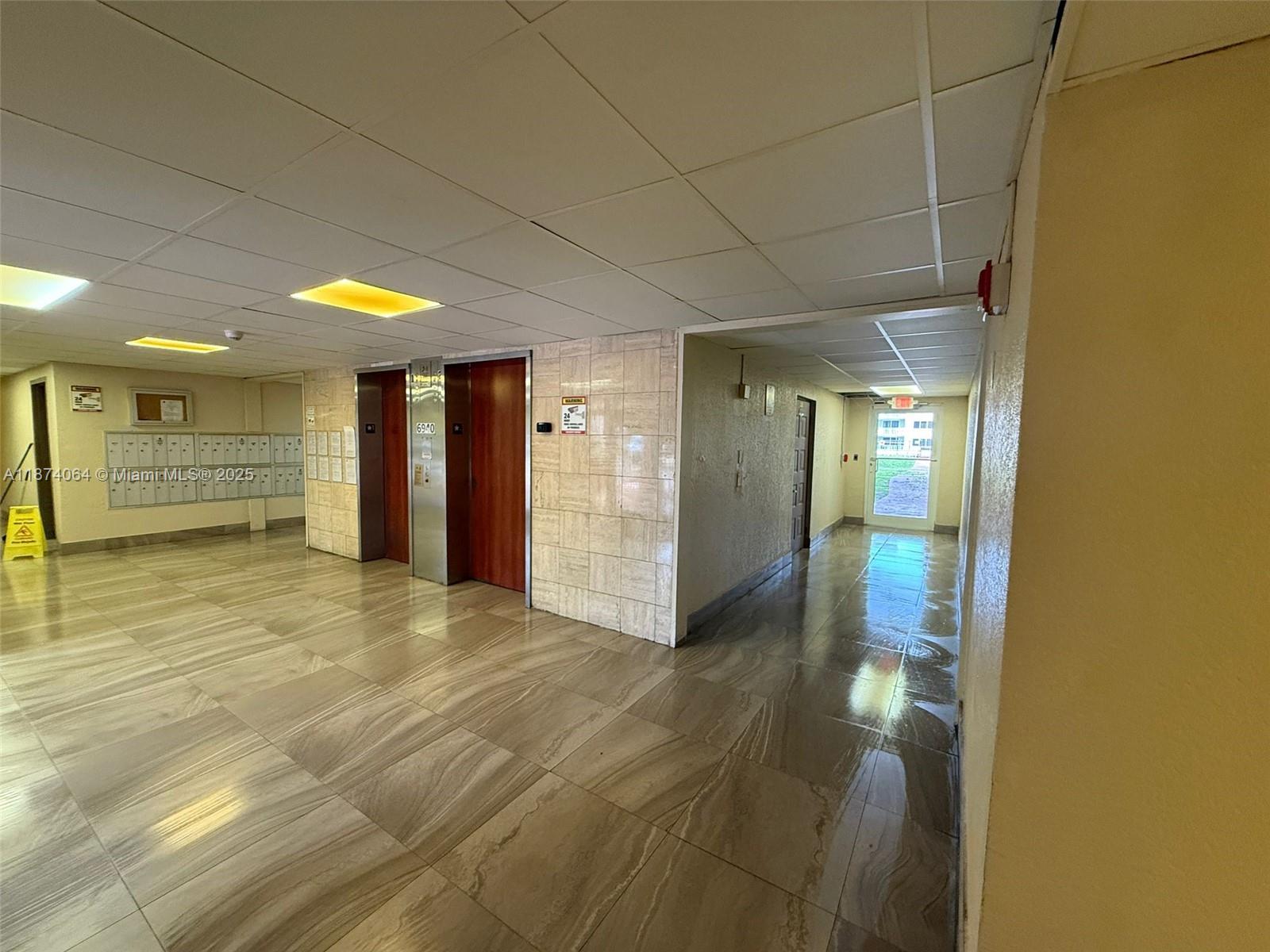 An apartment building lobby features tile flooring, two elevators, and a hallway leading to an exit — 6940 Miami Gardens Dr # 122, Hialeah FL 33015, HIALEAH, FL